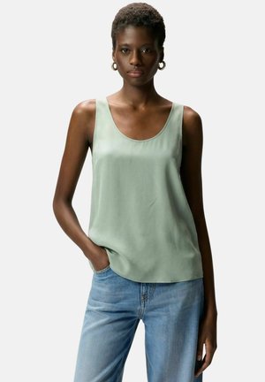 Giovane donna di colore che indossa orecchini a cerchio dorati, un top verde chiaro senza maniche e jeans blu, in piedi con una mano in tasca.