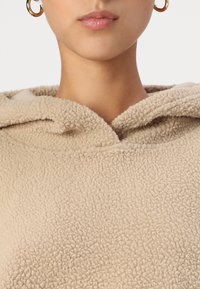 Sweat à capuche en polaire beige avec une surface texturée, comportant une poche avant et un capuchon avec cordon de serrage. Boucles d'oreilles en or visibles.