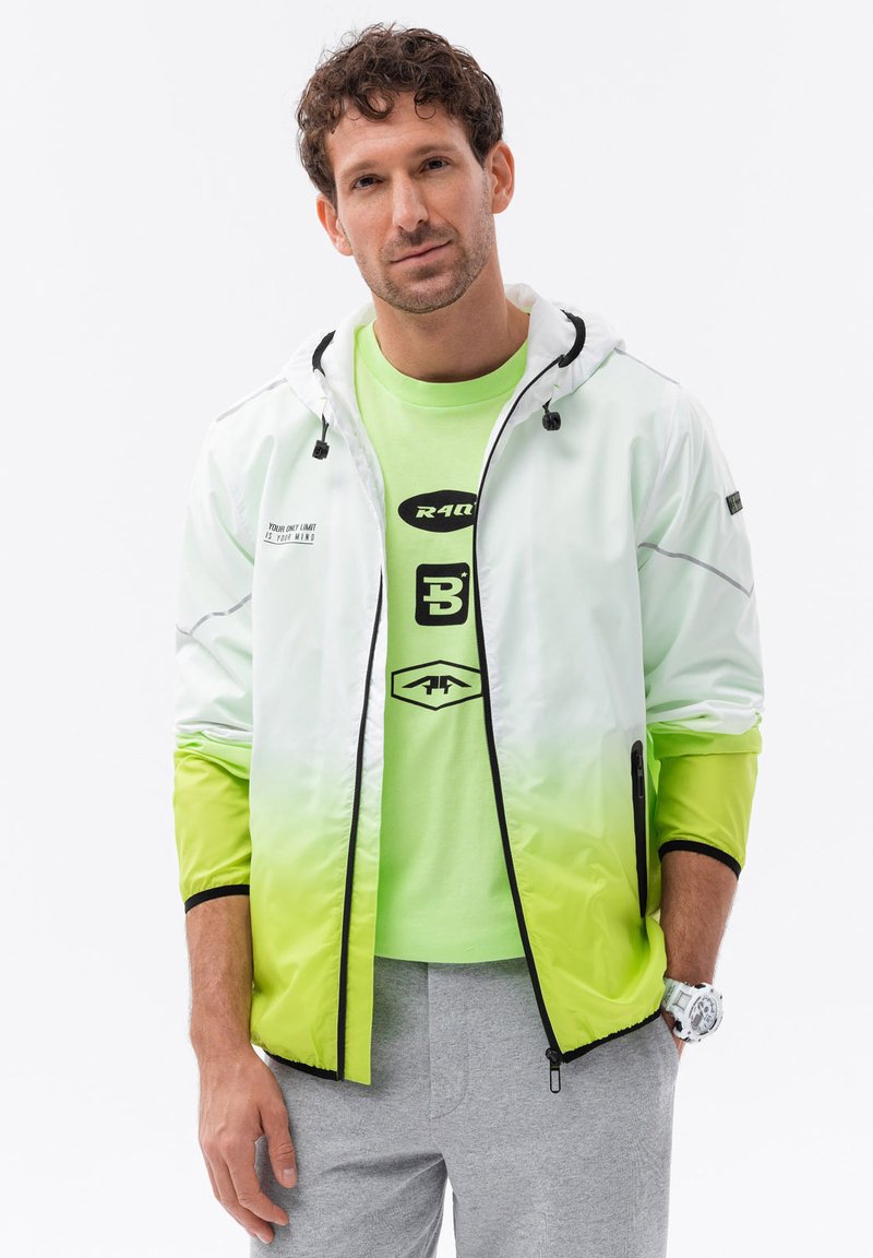 Giacca con gradiente bianco e lime, con cappuccio, tasche con zip e dettagli neri; indossata sopra una maglietta verde brillante e pantaloni della tuta grigi.