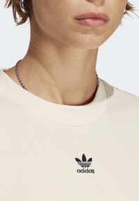 Cremefärgad sweatshirt i mjukt tyg, med ett ribbat rundt nacke och en liten svart Adidas-logga broderad på bröstet.