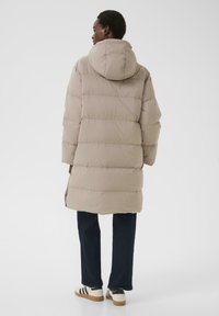 Bežna pufasta jakna s kapuljačom, ki ima vodoravne prešite dele. Model nosi temne kavbojke in bele teniske s črnimi detajli.