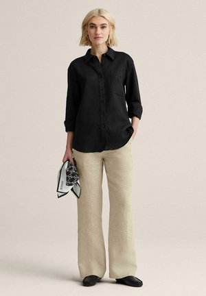 Kvinde iført en sort skjorte med knapper og beige vidde bukser, der holder et sort-hvidt mønstret tørklæde i venstre hånd.