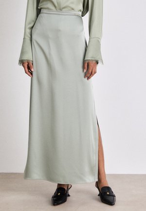 Mujer vistiendo una falda midi de seda verde salvia con abertura lateral, blusa a juego con puños con flecos y mocasines negros de tacón con detalle dorado.