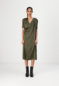 Olīvu zaļš midi kleita ar piekārtu priekšpusi, īsām piedurknēm un V veida kakla izgriezumu, kombinēta ar melnām potīšu zābakiem. Gluda tekstūra un viegla materiāla.