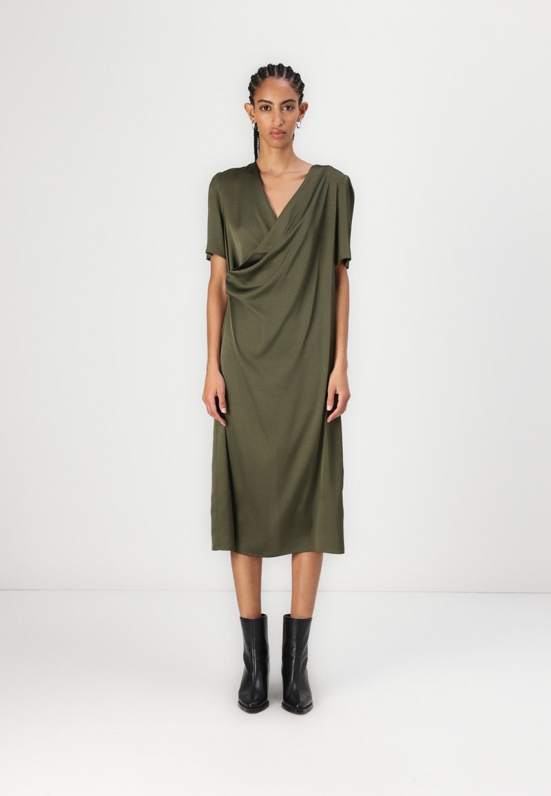 Olīvu zaļš midi kleita ar piekārtu priekšpusi, īsām piedurknēm un V veida kakla izgriezumu, kombinēta ar melnām potīšu zābakiem. Gluda tekstūra un viegla materiāla.