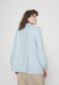 Blazer ajustado azul claro com mangas longas e ligeiramente volumosas, e um único ventos nas costas. O tecido tem uma textura suave, ideal para sobreposições.
