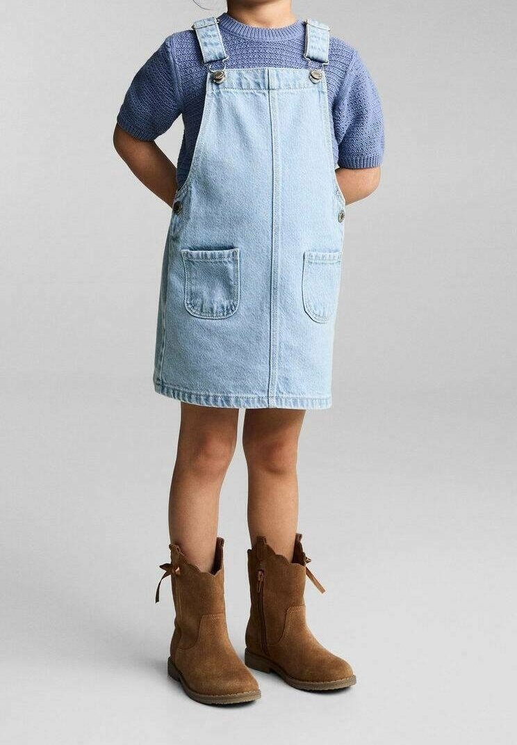 Enfant portant une robe salopette en jean bleu clair, un haut en maille violet à manches courtes et des bottes en suède marron, debout les mains derrière le dos.