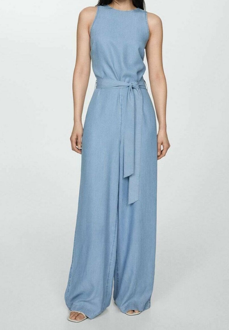 Femme portant une combinaison sans manches bleu clair à jambes larges avec une ceinture en tissu nouée à la taille, debout devant un fond uni.