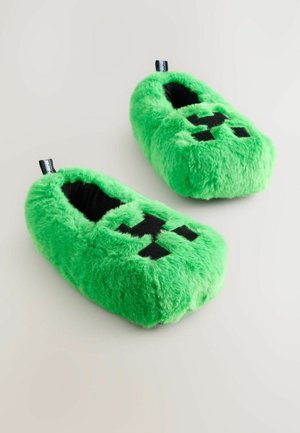 Chaussons en peluche verts avec une texture douce, une forme arrondie et un design de visage pixelisé noir à l'avant. L'intérieur est noir.