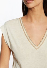 Light beige V-neck top made of soft fabric, featuring short sleeves and a decorative gold trim around the neckline.