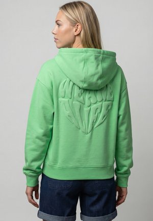 Blonde Frau mit hellgrünem Hoodie, die Kapuze aufgesetzt, mit dem aufgedruckten Text "WE ARE ALIVE" auf dem Rücken, kombiniert mit hochgekrempelten dunkelblauen Jeansshorts.