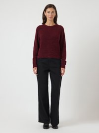 Jovem mulher em pé, a usar um suéter bordeaux, calças largas pretas, sapatos pretos e brincos de argola dourados, diante de um fundo branco plano.