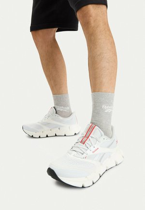 Piernas con pantalones cortos negros, calcetines grises de Reebok y zapatos deportivos blancos de Reebok con suelas gruesas y detalles en rojo sobre un fondo blanco.