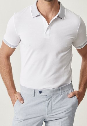 Mand iført en tætsiddende hvid polo med grå kant på krave og ærmer, kombineret med lyseblå teksturerede bukser, hænderne i lommerne.