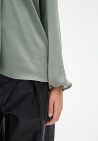 Top verde chiaro a maniche lunghe con vestibilità morbida e polsini arricciati, abbinato a pantaloni in pelle neri a vita alta. Tessuti lisci, dettagli minimi.