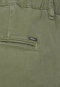 Närbild av olivgrön tyg som visar en sydd ficka med en liten metalltagg märkt "Pepe Jeans" nära den övre sömmen.