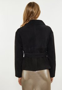 Schwarze Wildlederjacke mit breitem Kragen, tailliertem Schnitt und langen Ärmeln. Die Rückseite hat eine glatte Textur und ein dezentes Peplum-Design.