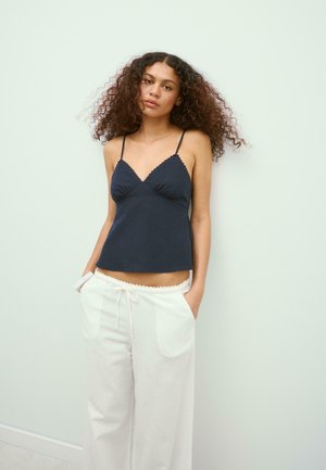 Jeune femme aux cheveux bouclés portant un haut sans manches bleu marine foncé et un pantalon large blanc à cordon, debout contre un mur pâle.