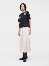 Femme portant un haut noir à manches courtes avec des motifs blancs, une longue jupe blanche avec des détails en dentelle, et des bottes noires mi-mollet, debout devant un fond uni.
