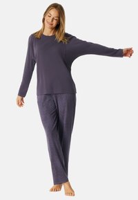 Lila Langarmoberteil mit lockerer Passform; passende lila Hose mit wellenförmig strukturiertem Muster. Weicher Stoff. Modell steht barfuß mit ausgestrecktem Arm.