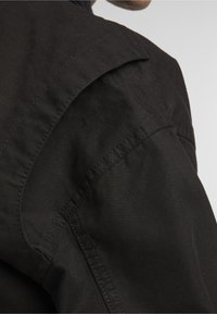 Czarna kurtka z teksturowanego materiału, z karczkiem na ramionach i szczegółowym przeszyciem wzdłuż szwów. Zbliżenie na obszar ramienia.