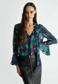 Blusa trasparente color teal con motivi astratti rosa e verdi, con bottone frontale, maniche lunghe con polsini a campana, indossata sopra una gonna nera con zip.