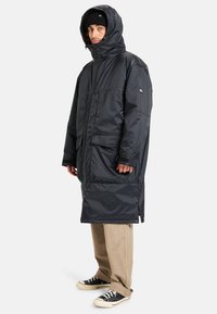 Zwarte gewatteerde lange jas met een capuchon, voorzien van een voorkant rits, twee grote voorzakken en elastische manchetten. Gemaakt met beige broek en zwarte schoenen.