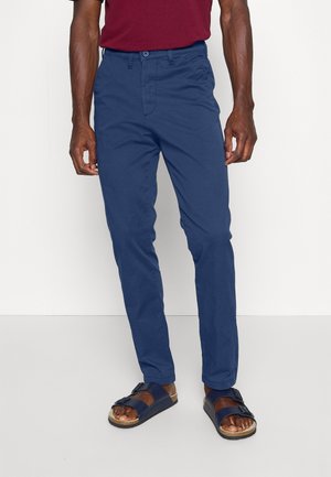 Marineblaue Baumwollhose mit geradem Schnitt, Fronttaschen und Knopfverschluss, kombiniert mit braunen Sandalen mit schwarzen Riemen.