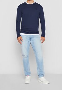 Navy long-sleeve shirt with white collar trim, paired with light blue jeans and white sneakers. Features small embroidered logo on chest.