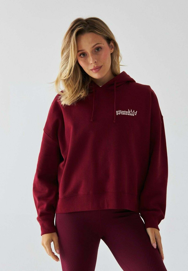Bordowa bluza z kapturem, z kieszenią z przodu, sznurkiem w kapturze oraz białym napisem "królewna pilates". Miękka tkanina i luźny krój.