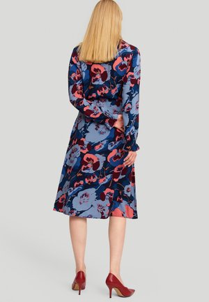Frau mit glattem blondem Haar, die ein blaues, pinkes und bordeauxfarbenes Blumenkleid und rote High Heels trägt, steht mit den Händen hinter dem Rücken.