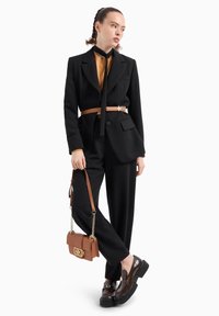 Completo nero su misura con cintura marrone, accessoriato con una borsa marrone e scarpe nere lucide. Presenta un dettaglio con logo dorato sulla borsa.