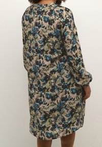 Abito floreale con motivi blu, neri e verdi su uno sfondo beige; presenta maniche lunghe e una vestibilità comoda.