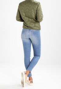 Olive green quilted jacket with a round collar, paired with fitted light blue jeans and silver-accented sneakers. Plain background.