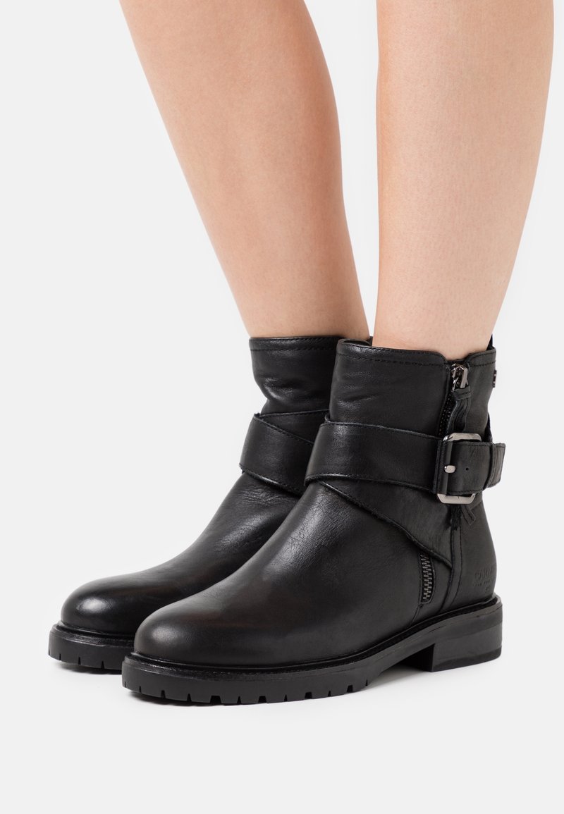 Black leather ankle boots with a rounded toe, side zipper, and a decorative strap with a metallic buckle. Textured rubber sole.
