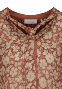 Bluse mit Blumenmuster in Pfirsichfarbe und weißen Blumen. Sie verfügt über einen Knotenverschluss am Hals und hat eine glatte Stoffstruktur. Hergestellt in Portugal.