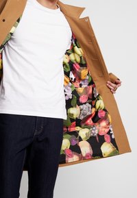 Personne portant un manteau camel montrant une doublure intérieure colorée à motif de fruits, associée à une chemise blanche et un jean foncé.