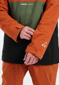 Farbenblockierte Jacke mit einem orangefarbenen und grünen oberen Teil und einem schwarzen unteren Abschnitt. Mit Klettverschluss an den Manschetten, Branding und einer seitlichen Reißverschlusstasche.