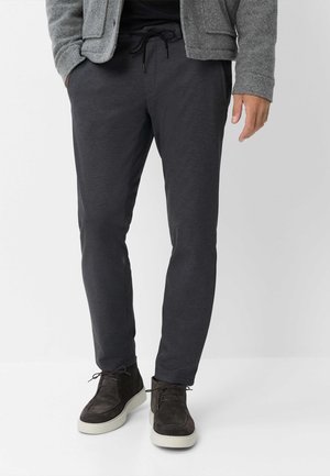 Uomo con pantaloni grigio scuro con coulisse, giacca grigia e stivali in camoscio marrone scuro su sfondo chiaro uniforme.