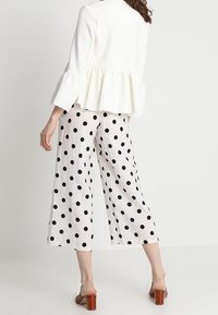 Biała kurtka peplum z falbaniastymi rękawami połączona z czarnymi spodniami culottes w kropki. Brązowe buty na słupku. Jasne tło.