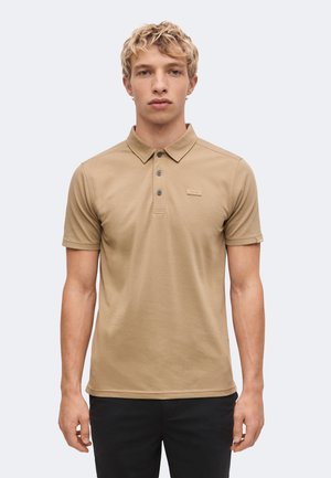 Ung mand med kort krøllet blondt hår iført en beige poloshirt og sorte bukser står mod en ensfarvet lys baggrund.