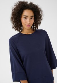 Námornícky modrý mikina s okrúhlym výstrihom, rukávmi 3/4 raglan a mäkkou, hladkou štruktúrou. Má voľný strih a minimálne švy.