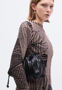 Sac en cuir noir à plis avec une fermeture à cordon, tenu contre un vêtement à manches longues à motifs aux textures rose et noir.