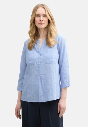 Light blue, long-sleeve blouse with a V-neck, two front pockets, and a textured fabric featuring a subtle pattern. Relaxed fit.