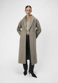 Largo abrigo de lana gris con solapa ancha, cierre de un solo botón y bolsillos. Llevado sobre una blusa beige y jeans negros, combinado con tacones negros.