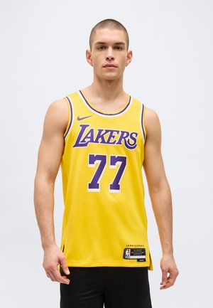 Muž v žltom basketbalovom drese Los Angeles Lakers s číslom 77, stojaci na jednoduchom svetlom pozadí.