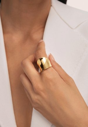 Gold-plattierter Ring mit einer glatten, polierten Oberfläche, der eine breite Bandbreite und ein sanft geschwungenes Design aufweist, getragen an einer Hand vor einem weißen Hintergrund.