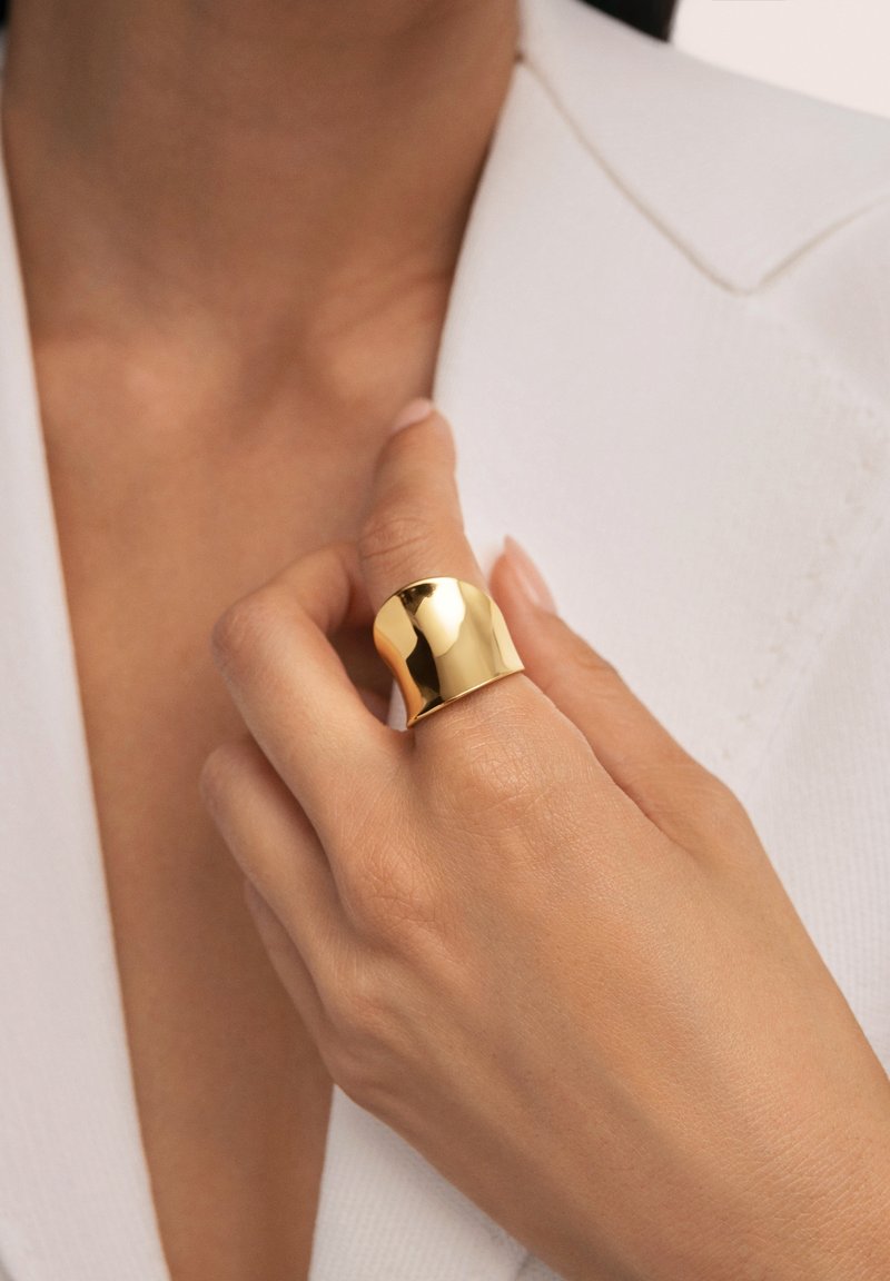 Anello placcato oro con superficie liscia e lucida, caratterizzato da una fascia larga e un design morbido e curvo, indossato su una mano su sfondo bianco.