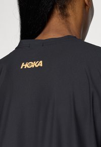 Čierne športové tričko s textúrovanou, vlhkosť odvádzajúcou tkaninou a výrazným zlatým logom "HOKA" na hornej časti chrbta.