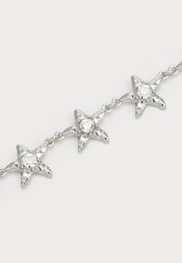 Sølvstjernesmykker med klare runde ædelsten forbundet med små kæder på en ensfarvet lys baggrund.